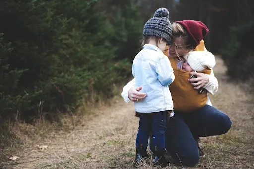 Anxious Parenting: How To Deal With Anxiety As A Stressed-Out Single Mum 4 Mother babywearing small baby on her chest, bending down hugging a toddler in the park